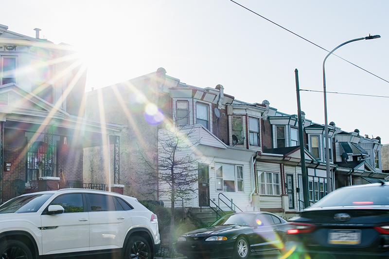A photograph of Philadelphia row homes. We can see some cars in the street, and a bright lens flare breaks over the houses where the sun has risen.
