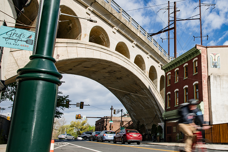 Photo taken on a sunny day of the Manyunk Bridge, in Manayunk, Philadelphia. Photo looks up from a low-angle to include street with cars and cyclist, underside of bridge, and blue sky with light clouds.
