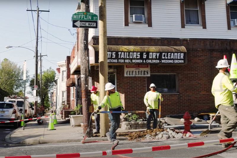 A photograph featuring four male construction workers, in hard hats and yellow vests, working on Mifflin St, in Philadelphia.